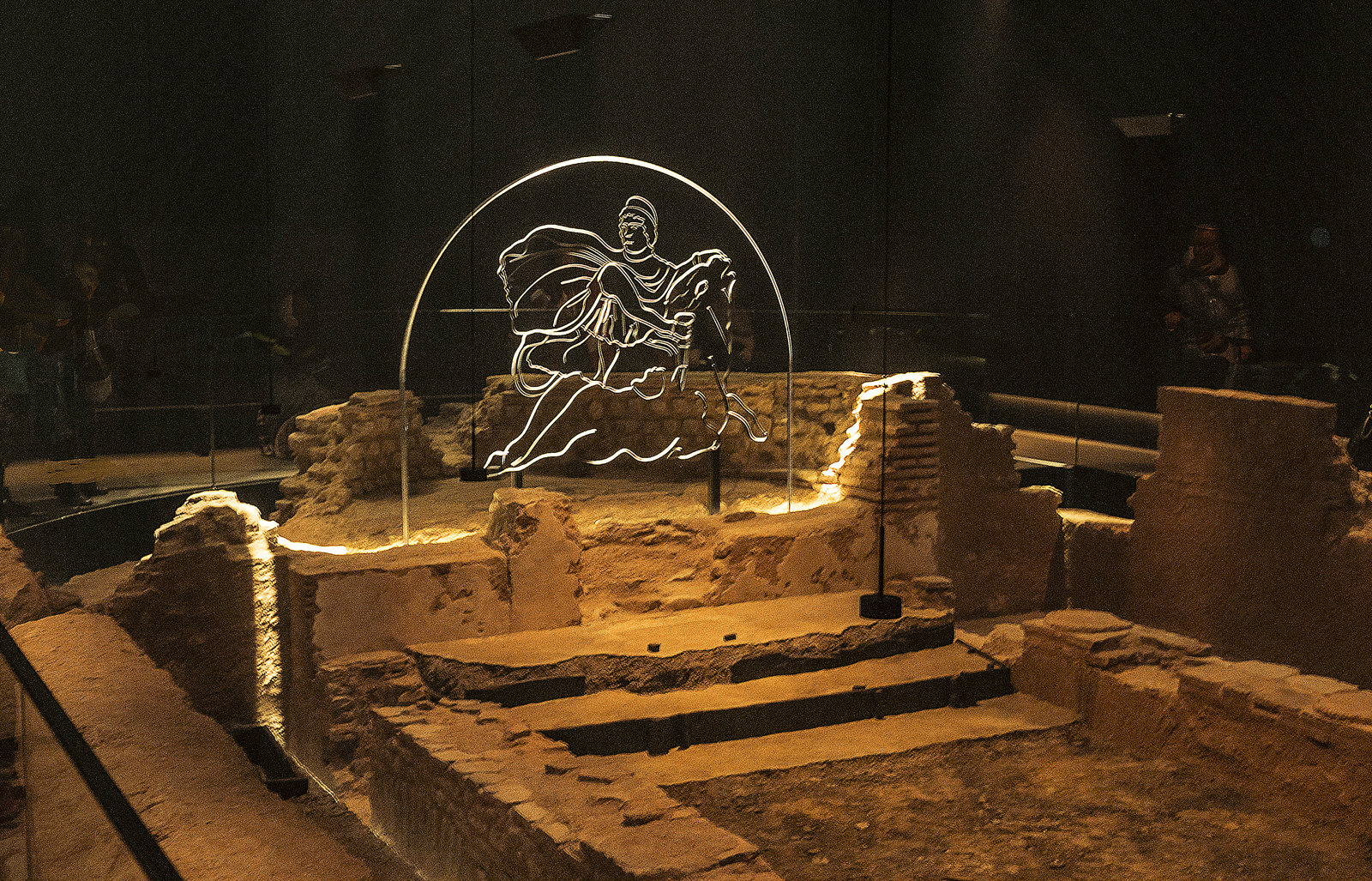 Interior of the reconstructed London Mithraeum below Bloomberg's headquarters: low stone temple walls lit in haze, seven metres beneath the modern street