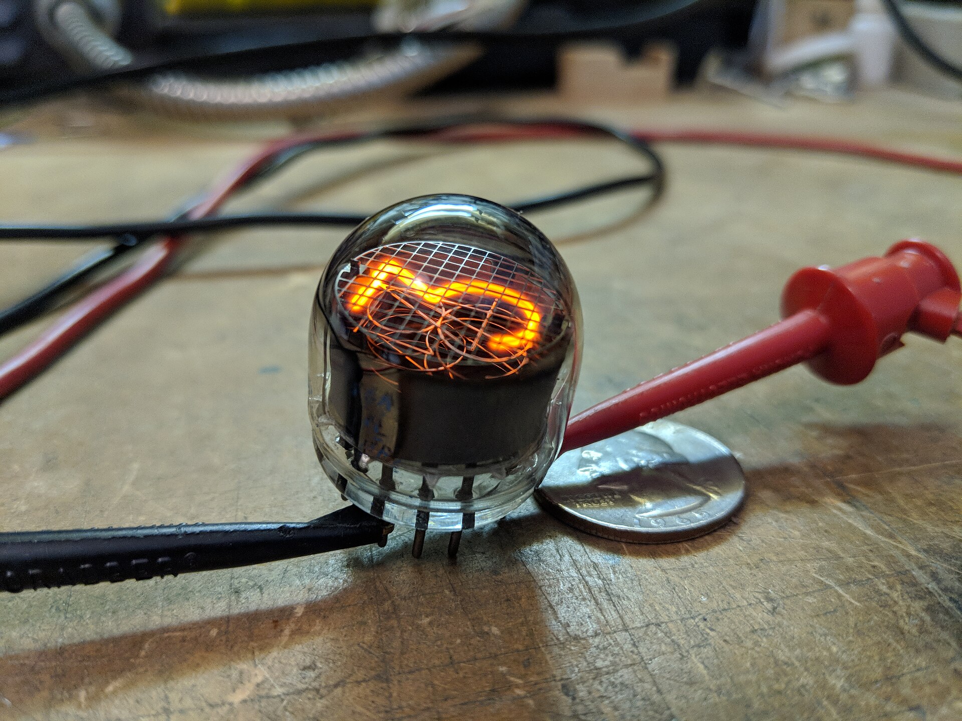 A glowing Nixie tube wired to test leads, shown beside a US quarter for scale