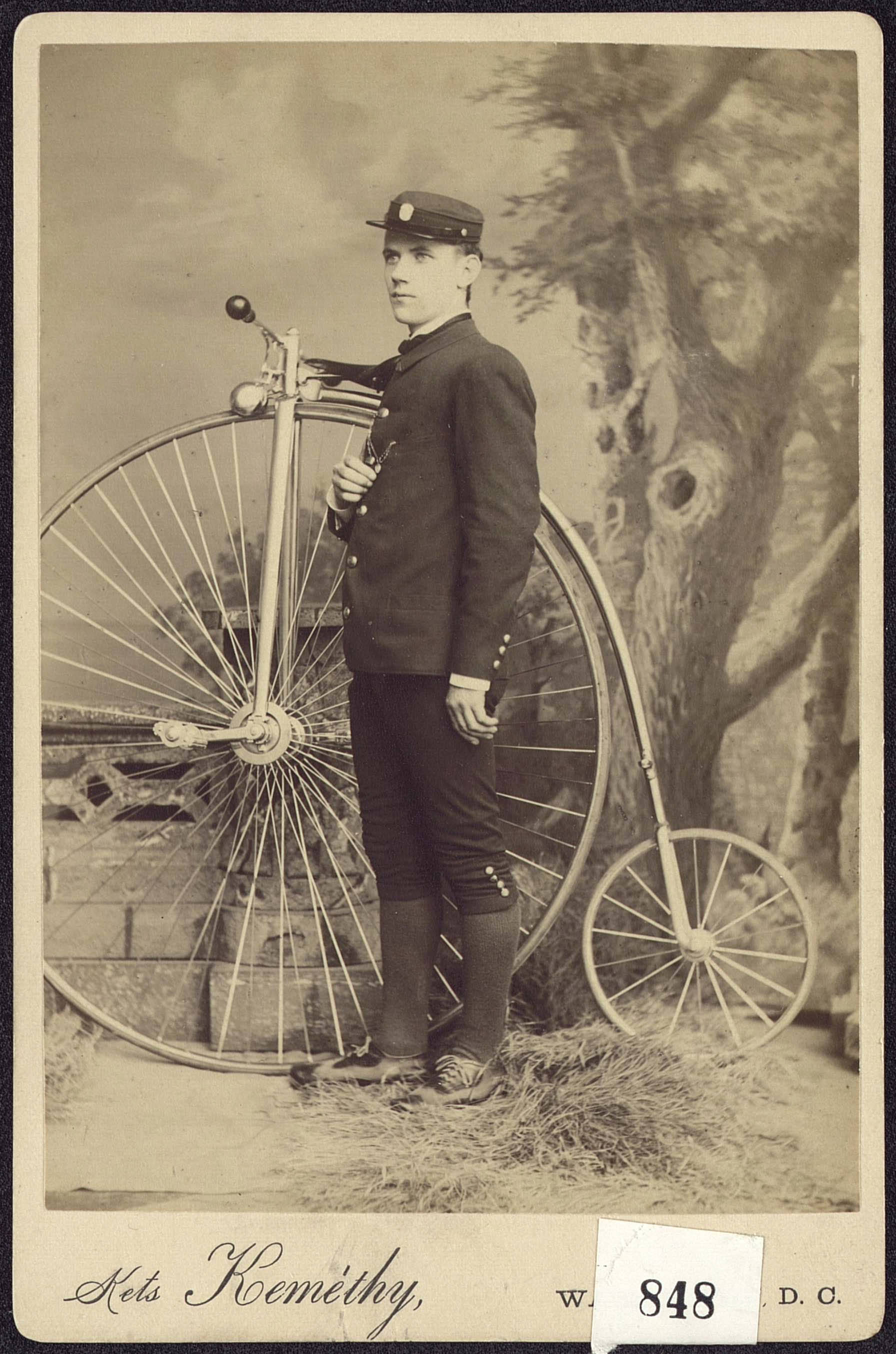 Late-19th-century portrait photograph of a man standing beside a penny-farthing bicycle with its tall front wheel